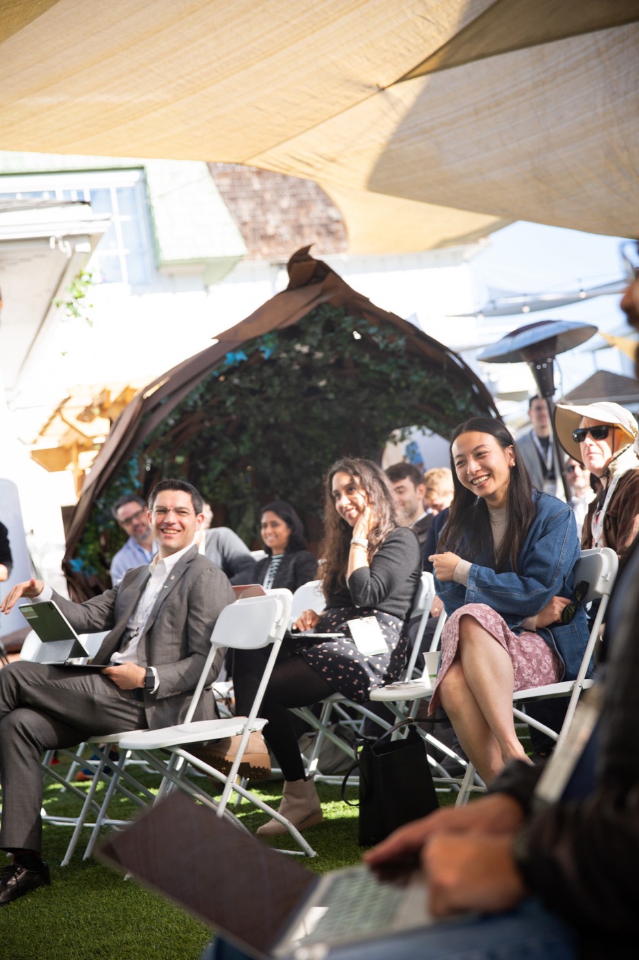 Attendees on folding chairs under a shade sail at an outdoor session, laughing and smiling.