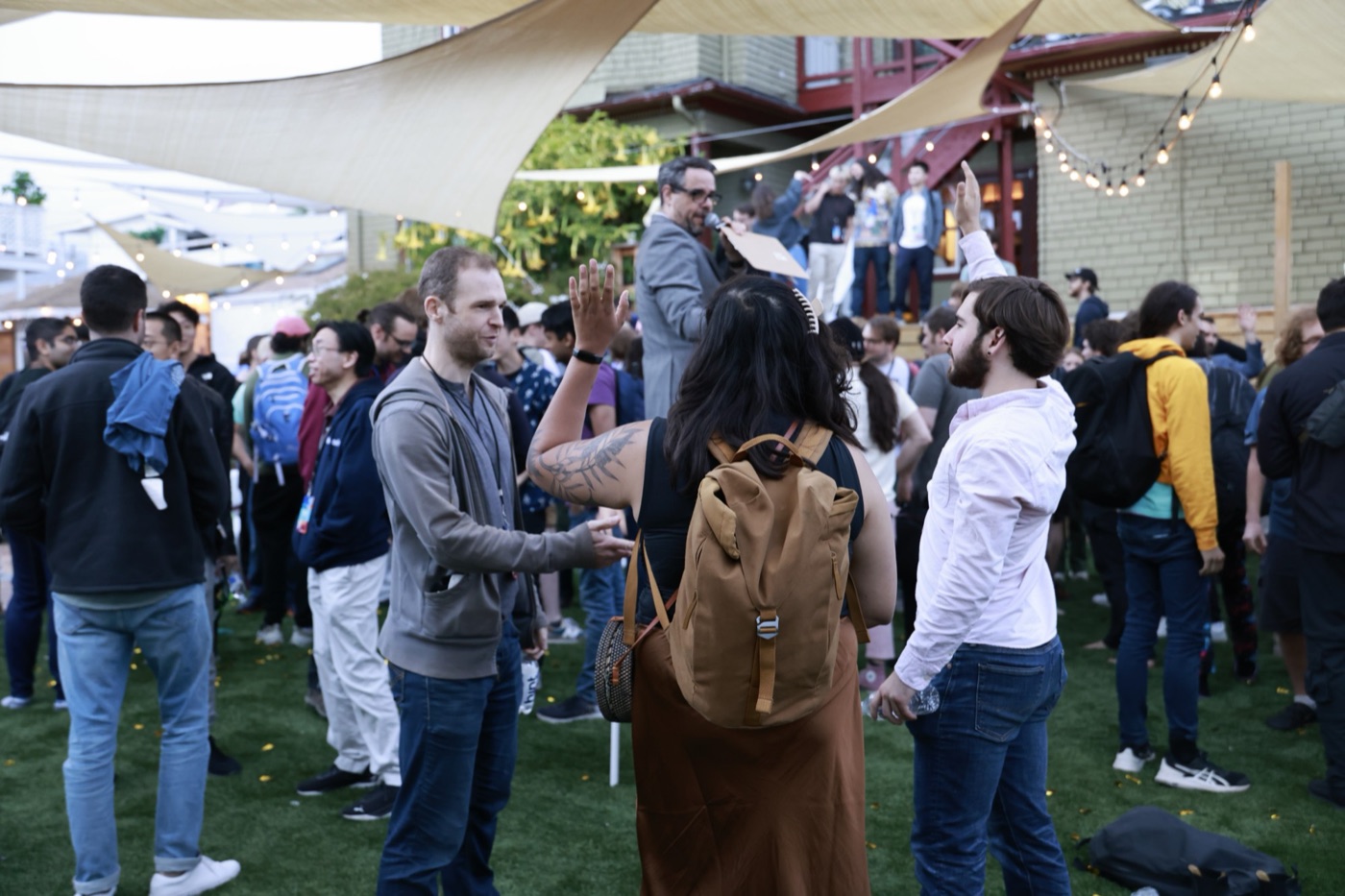 Misha with a microphone directing an outdoor crowd, as attendees in the foreground raise their hands and talk in pairs.