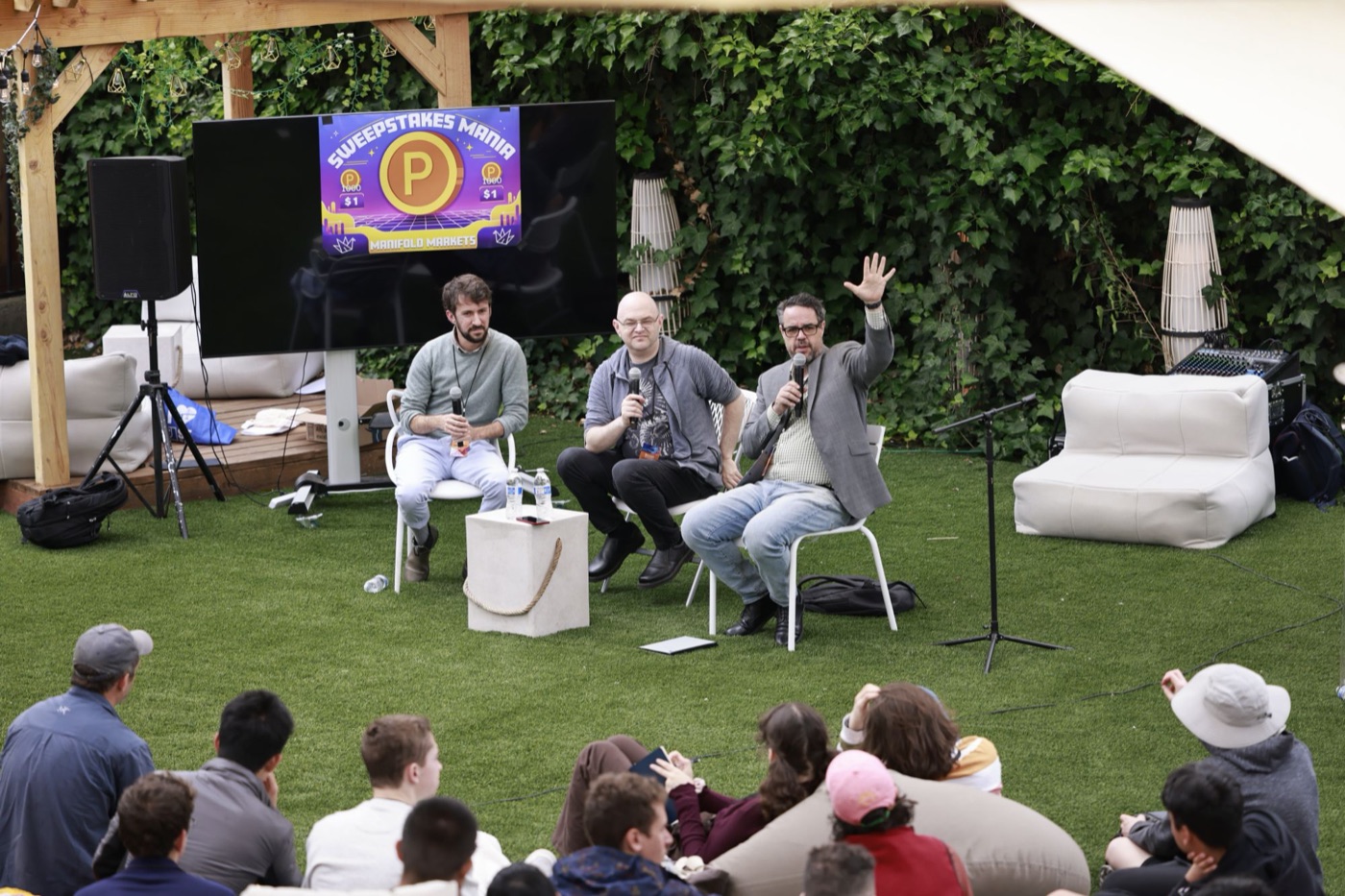 Misha raising a hand as he speaks on an outdoor panel, with an audience seated on the lawn in front.